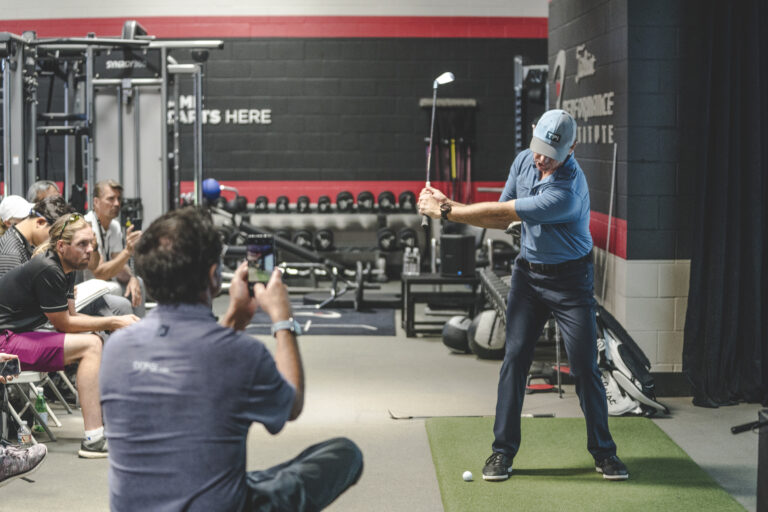 Golf instructor demonstrating swing technique indoors to a group of students in a training facility